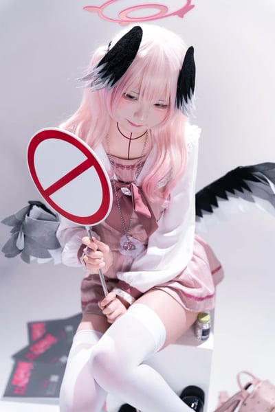 这是一张ACG cosplay写真图，coser完美还原了手游《蔚蓝档案》中的角色下江小春：粉色长发搭配黑灰渐变的翅膀耳饰，头顶还有后期添加的粉色光环，身穿粉白相间的可爱洛丽塔风格服饰，搭配白色过膝袜。她双手握着一个禁止通行的圆形标识牌，面带软萌羞涩的微笑，乖巧地坐着，整体氛围甜美治愈，尽显角色的软萌气质。