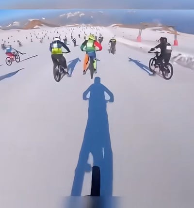 画面展现了一片广阔的雪山赛道，众多骑手正骑着自行车从斜坡上疾驰而下。前景中清晰可见拍摄者的影子，其双手叉腰，仿佛在注视着这场激烈的角逐。远处的雪山连绵起伏，天空湛蓝，阳光明媚，整个场景充满了动感与冬季户外运动的活力。