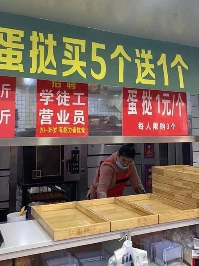 这是一家街边烘焙店的柜台实拍图，上方绿色招牌标注「蛋挞买5个送1个」，但右侧红色海报却写着「蛋挞1元/个 每人限购3个」，同时柜台还张贴着招聘学徒工和营业员的启事，要求年龄20-35岁、有能力者优先。柜台内的工作人员正低头整理木质托盘。<br>矛盾点：买5送1的促销活动和每人限购3个的规则完全冲突，属于商家促销信息设置不合理的搞笑场景，让人无法正常参与活动。