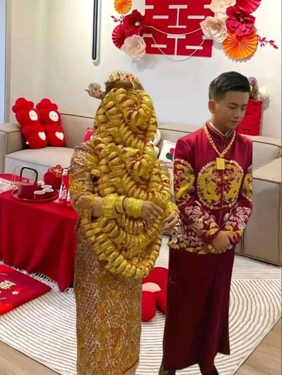 这是一场充满中式传统氛围的婚礼现场，新娘身着金色礼服，全身被大量黄金手镯、项链等首饰挂满，数量多到几乎覆盖上半身，视觉冲击力极强；新郎则穿着绣有龙凤纹样的红色中式礼服，佩戴着黄金项链与金牌。背景布置以喜庆的红色为主，墙面装饰着大红喜字与纸艺花饰，沙发旁的桌案上摆放着中式茶具、可口可乐与喜庆摆件，整体氛围隆重奢华。这种大量佩戴黄金的形式是福建、广东等地的婚礼习俗，是彰显嫁妆丰厚的传统方式。