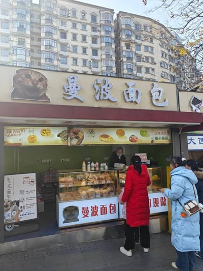 这是一张街头面包店的实拍照片，展示了名为“曼波面包”的店铺场景。店铺门头上方有“曼波面包”四个大字，左侧配有一个猫咪头像的标志。店面前方的展示柜里摆放着各种面包糕点，一名店员正在柜台后为顾客服务。几位穿着冬季外套（红色和蓝色羽绒服）的顾客正在排队购买，背景是一栋多层居民楼，路边有带有黄叶的树木，整体呈现出温馨的城市日常氛围。店铺玻璃上贴有“新店尝鲜”“现烤现卖 绝不隔夜”等促销字样，下方还有“曼波面包 现做”的招牌。