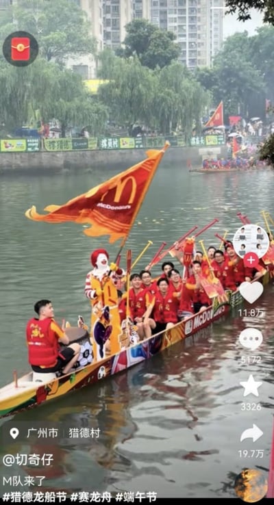 这是一张竖屏的社交媒体短视频截图，拍摄地点为广州市猎德村的河道，场景是端午节期间举办的猎德龙舟节活动。画面主体是一艘获得麦当劳赞助的参赛龙舟，船身装饰有麦当劳品牌元素，船头立着麦当劳叔叔的人形立牌，还插着一面印有麦当劳金色M标志和品牌名的橙色旗帜，龙舟上的参赛队员都穿着统一的红色队服，船头位置有一名鼓手负责击鼓控速，其余队员手持船桨坐在船身，全员神态轻松愉悦，士气高昂。背景可见河道两岸的垂柳、高层居民楼，还有观赛的人群和其他参赛的龙舟，整体充满端午民俗活动的热闹氛围。截图保留了短视频平台的交互界面元素：左上角有红包悬浮按钮，右侧依次是点赞、评论、收藏、转发的功能按钮及对应数据，左下角标注了定位、发布者账号、配文以及相关话题标签。