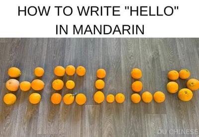 图片上方是黑色英文标题'HOW TO WRITE "HELLO" IN MANDARIN'，下方木质地板上用多个橘子排列组成中文'你好'二字，创意展示如何用中文书写'hello'。整体风格轻松有趣，适合语言学习场景使用。