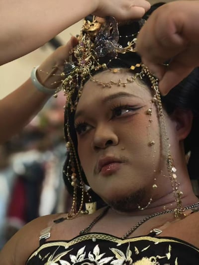 画面中一位留有胡须的男士，正被他人协助佩戴一款装饰着大量金链、珍珠的华丽古风头冠。男士化着精致的上扬眼线古风妆容，眼尾上挑，搭配浓密睫毛，和自身硬朗的外形形成强烈的反差感，他抬头看向头冠方向，神情平静中带着一丝期待，整体场景充满趣味和独特的创意。