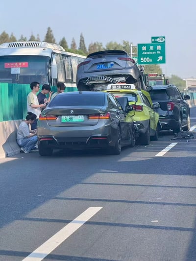上海某高速公路上发生的多车连环相撞事故现场。画面中可见一辆银色宝马轿车(沪A·BK)与一辆黄色轿车发生碰撞，更令人震惊的是一辆强生出租车被叠压在宝马车顶部。事故车辆停在道路右侧，几名涉事人员站在护栏旁，其中一人蹲在地上查看情况。背景中可见一辆标有'工人先锋号'的公交车和道路指示牌显示距离剑川路还有500米。