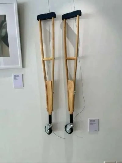 图片展示了两根挂在白色墙壁上的木制拐杖，每根拐杖底部都被加装了一个小型轮子。拐杖顶部为黑色软垫，中间有横向支撑结构，整体呈自然木色。左侧墙壁上挂着一幅带框的装饰画，拐杖下方各有一张白色小卡片（可能是说明文字）。这一“发明”据称旨在解决残疾人拄拐杖步行缓慢的问题，但从实际使用逻辑来看，拐杖依赖支撑与交替抬升动作，加装轮子后可能导致稳定性丧失，反而存在安全隐患，呈现出一种荒诞的幽默感。