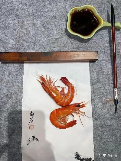 画面中央铺展着一张白纸，纸上摆放两只鲜红蜷曲的熟虾，姿态宛如国画中的生灵。纸左下角有黑色毛笔题字“白石”及红色印章，右下角点缀水墨笔触小图。白纸上方压着木质镇纸，右侧依次为浅绿色酱汁碟（盛深色液体）与红杆毛笔，背景为灰色布艺。整体以真实熟虾模仿齐白石水墨虾图，搭配文房元素，形成传统艺术与美食结合的创意场景。