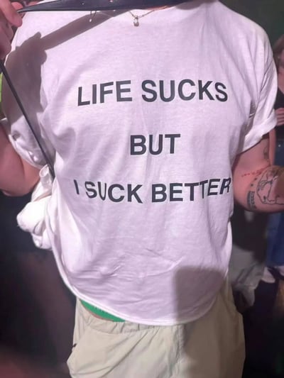 图片展示了一个人背影，穿着印有黑色大写文字“LIFE SUCKS BUT I SUCK BETTER”的白色T恤。此人左臂有纹身，手持黑色物体靠近肩膀，背景为昏暗的社交场合环境。文字通过自嘲的方式表达了在生活不如意时的乐观态度，带有黑色幽默色彩。