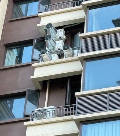图片展示了一栋多层居民楼的外立面，建筑主体为棕色与米白色相间。中间楼层的一个阳台位置密集安装了四个银色金属排气扇，排气扇呈倾斜状向外伸出，部分窗户处于打开状态，可看到室内窗帘。整体场景为白天自然光拍摄，窗户玻璃反射出天空景象，未见人物活动。