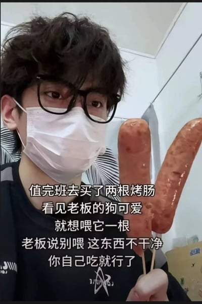这是一张真人实拍的图文混排静态图，画面主体是一位留着卷发、佩戴黑框眼镜与白色口罩的年轻男性，他身着黑色上衣，手里举着两根油亮的烤肠看向镜头。图片上叠加了白色的段子文案，内容为值完班去买了两根烤肠，看见老板的狗可爱，就想喂它一根，老板说别喂 这东西不干净，你自己吃就行了。文案通过老板“烤肠不干净不让喂狗却让当事人自己吃”的表述，形成了反向讽刺的搞笑效果，暗含调侃老板不把员工健康当回事、员工地位不如老板的狗的趣味梗点。