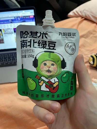 图片中一只手握着一袋九阳豆浆的「哈基米南北绿豆」豆浆植物饮料，包装袋设计充满趣味。正面印有一只戴着绿色绿豆造型头套、戴着黑色耳机的卡通猫咪，猫咪咧嘴笑着，双手抱着红色吉他，显得十分活泼可爱。包装上标注了产品名称「哈基米 南北绿豆」、净含量150g、品牌「九阳豆浆 Joyoung Soymilk」以及「鲜磨」「0脂肪」等信息，底部还有标语「哈基米的音乐才是真正的音乐」。背景中可见一台打开的笔记本电脑和铺着浅色床单的床铺，营造出居家休闲的场景。

图片结合了网络热词「哈基米」（源自对猫咪的可爱称呼）作为产品名称，将流行文化元素融入食品包装设计，具有明显的 meme 营销特征，让普通食品包装变得更具话题性和趣味性。