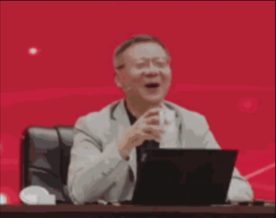 这是一段动态GIF，画面中是学者金灿荣坐在会议桌后，面前摆放着笔记本电脑，他原本状态平稳，突然露出夸张的惊讶表情，同时举起手中的水杯，动作和表情极具戏剧性，背景是红色的会场背景板，这个片段常被用来制作表情包，表达惊讶、兴奋、意外等强烈情绪。