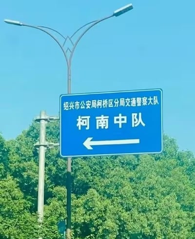 图片展示了一块安装在路灯杆上的蓝色交通指示牌，牌面文字为“绍兴市公安局柯桥区分局交通警察大队 柯南中队”，下方配有向左的白色箭头。背景是茂密的绿树和晴朗的蓝天，整体画面清晰自然。

该路牌因“柯南中队”的名称产生趣味联想——“柯南”与日本著名侦探动漫《名侦探柯南》主角同名，使严肃的交警中队名称带有轻松幽默的效果，成为网友关注的亮点。