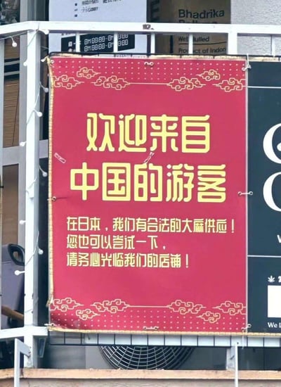 这是一张在日本拍摄的店铺宣传标语牌照片，标语牌为红色底搭配黄色字体，带有中式云纹装饰边框，主标语为“欢迎来自中国的游客”，下方文字称在日本有合法的大麻供应，邀请中国游客尝试并光临店铺。需要注意的是，大麻在中国属于违禁品，该标语针对中国游客宣传大麻，存在明显的不良引导，违背中国的法律法规和公序良俗。