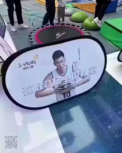 这是一张儿童运动场馆内的实拍照片，前景是一块椭圆形带黑色边框的品牌宣传广告牌，牌面印有篮球运动员郭艾伦的形象，搭配“Joykids 卓跃儿童”的品牌标识与“强强引领儿童”的宣传语，还有竖排的“品牌代言人 郭艾伦”字样，同时牌面带有“分享群267694793”的水印文字。背景可见ADKING品牌的蹦床、绿色运动软垫等儿童体能训练设施，还有几位人员在场馆内活动，整体呈现出儿童专业体能训练机构的场景氛围。