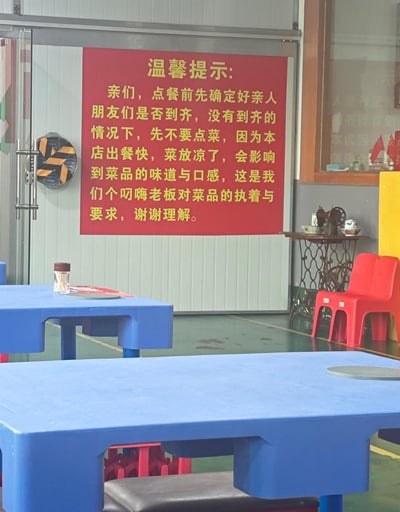 这是一张实拍的餐馆店内场景照片，画面前景是两张蓝色的连体餐桌，近处蓝色餐桌下方露出带灰色软垫的座位，左侧稍远的蓝色餐桌上放置了一个透明的牙签容器；背景的浅色玻璃隔断门上贴着一块醒目的红底黄字温馨提示牌，提示牌内容告知顾客：点餐需要等亲友全部到齐再点，人没到齐不要提前点菜，因为本店出餐速度很快，提前点的菜会放凉影响味道和口感，这是店主对菜品品质的执着要求，恳请顾客理解。提示牌左侧是玻璃门的金属门把手，旁侧挂有一个拼色装饰盘；提示牌右侧的墙边摆放了一张镂空铁艺边桌，桌上放置了老式缝纫机模型和装饰瓷碗，边桌旁靠墙放着一把红色塑料靠背椅，整体是真实的餐馆用餐区场景。