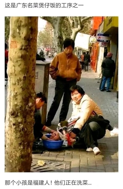 这是一张利用视觉错位结合网络地域梗制造搞笑效果的图片。画面是真实的街头场景：一位大人蹲在地上抱着小孩，小孩的头部伸进蓝色塑料盆中，旁边还有一人在盆边，从拍摄角度看，视觉上形成了“在洗小孩”的错觉；图片上下配有玩梗文字，上方写“这是广东名菜煲仔饭的工序之一”，下方写“那个小孩是福建人！他们正在洗菜...”，结合网络上“广东人吃福建人”的趣味玩笑梗，以及“煲仔饭”的谐音，制造出荒诞的喜剧效果，实际场景是大人在给小孩清洁脸部，只是拍摄角度造成了错位错觉。