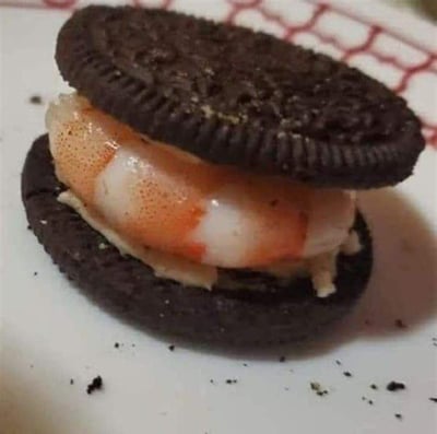 这是一张静态实拍的单图，展示了一款极具违和感的混搭食物：将奥利奥饼干的原有夹心移除，中间夹入了一只去壳的熟虾仁，虾仁下方还残留有少量类似酱料的物质，组合成了一个不伦不类的“夹心饼干”。甜口的奥利奥与咸鲜的虾仁进行混搭，完全打破了常规的饮食搭配逻辑，属于典型的网络整活黑暗料理，视觉上充满了猎奇感和搞笑的反差效果。