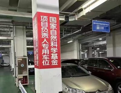 室内停车场场景，中央白色立柱上悬挂红色竖幅标语，内容为“国家自然科学基金项目负责人专用车位”。周围停放多辆轿车，背景可见停车场常见的管线、照明设备及模糊的蓝色指示牌。整体环境整洁，标语醒目，反映特定身份人员的专用停车设施。