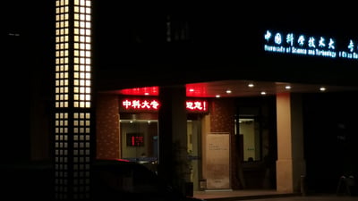 这是一张夜晚拍摄的中国科学技术大学校内某建筑入口的实拍照片。画面左侧是带有暖黄色灯光的格栅式建筑结构，入口上方亮着蓝白色的中英文校名牌，入口处的红色电子标语显示“中科大专 迎您！”，入口内透出室内灯光，门口立有一块展板，前景可见黑色车辆的局部，整体呈现出静谧温馨的校园夜晚氛围。