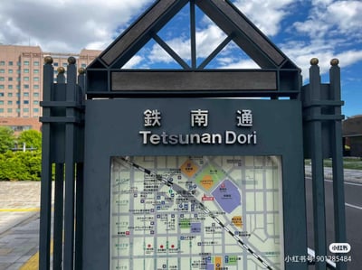 实拍日本街道的铁南通（Tetsunan Dori）指示牌，主体为黑色金属结构，顶部呈三角形镂空设计。牌面上方用中日双语标注名称，下方嵌有彩色区域地图，标注了JR带广职、酒店、商店等设施位置。背景可见红砖建筑、绿色灌木丛及蓝天白云，画面右下角有水印显示小红书用户信息。整体构图清晰，展现了典型的日式街道标识风格。
