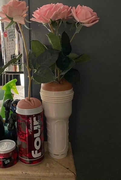 这张图片展示了一个创意DIY场景：三朵粉色玫瑰花被插在两个非常规容器中——一个红色的FOLK饮料罐和一个白色的手电筒。背景中可以看到一个绿色喷壶和一些小雕像，营造出一种生活气息浓厚的居家环境。这种将日常用品改造成花瓶的创意展现了废物利用的环保理念和生活小技巧。