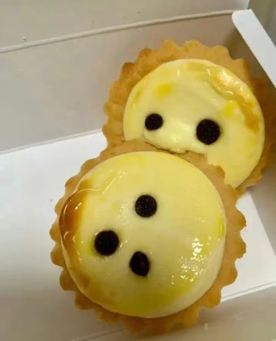 图片展示了两个芝士挞叠放在一起，并且表面上的巧克力豆排布巧合地形成了类似“呆脸”的表情包效果。其中一个挞的两颗巧克力豆像极了眼睛，被另一个叠在下方的芝士挞的三个巧克力豆构成了“鼻子”和“嘴巴”，整个场景带有强烈的戏谑感和拟人化幽默。由于点缀的位置和叠放方式，整体画面像是一个呆萌脸的表情，非常适合各种网络调侃或无语场合。

图片的有趣点在于食物被赋予了人物表情的效果，形成了一种强烈的“物人合一”的谐音梗（挞脸/塌脸），充满网络幽默气息。