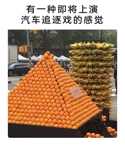 这是一张拍摄于国外街头市集的照片，商家为吸引顾客，用大量橙子堆叠出类似汽车车头与车身的造型，旁边用成串的香蕉堆成另一辆“汽车”的模样，从视觉效果上看如同两辆汽车停靠在路边。图片上方配文“有一种即将上演汽车追逐戏的感觉”，为这个创意水果造型增添了趣味氛围。背景中可以看到真实的行驶车辆、行人和路边的白色帐篷，整体是热闹的街头市集场景，这种创意的水果摆放方式充满巧思，十分吸睛。