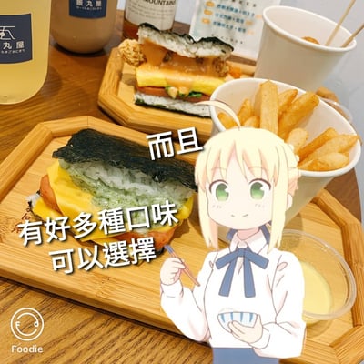 图片展示了一桌美味的日式快餐，包括两个不同口味的饭团三明治、一份薯条和几杯饮料。饭团三明治层次丰富，包含海苔、米饭、芝士、香肠和炸鸡等食材。图片右侧添加了一个金发绿眼的动漫少女形象，她手持筷子和碗，面带微笑，显得十分开心。图片上有文字标注："有好多種口味可以選擇 而且"，暗示这些食物有多种口味可供选择。