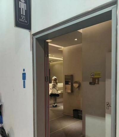 这是一张实拍的公共卫生间场景图，画面左侧墙面有蓝底男性标识和黑底白字的“男 MEN”男卫生间标牌，透过打开的男厕门，可以看到内部是整洁的米色调公共卫生间，配备洗手台、擦手纸盒、卷纸架等设施，墙上贴有“禁止吸烟 No Smoking”的标识。卫生间内的洗手区域站着一位穿着黑白女仆装、戴着白色头饰的金发coser，人物背对着镜头，和男卫生间的场景形成强烈的反差感，营造出趣味的视觉效果。