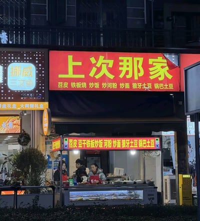 这是一张夜晚拍摄的街边小吃店实拍图，店铺的红色招牌用醒目的黄色大字写着极具记忆点的店名‘上次那家’，下方标注了苕皮、铁板烧、炒饭等多种小吃品类，店内有几位店员正在忙碌，旁边紧邻着一家‘挪威三文鱼’店铺。
这个店名采用口语化的表述，精准抓住了顾客“想找之前吃过的那家店”的消费心理，自带亲切感和记忆点，十分接地气又有趣。