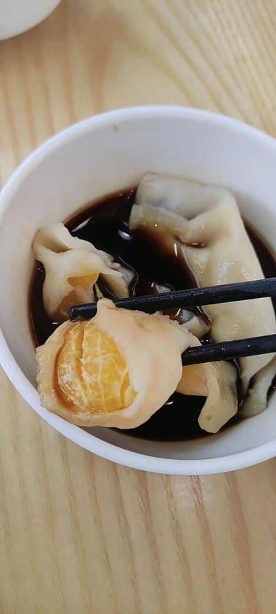 这张竖屏实拍照片里，白色小碗盛着深色蘸汁，里面泡着几个外形酷似饺子的食物。一双黑色筷子夹起其中一个，面皮中露出完整的橘子果肉，仿佛是把橘子当作馅料包进了饺子皮中，呈现出一种充满创意的新奇搭配，类似趣味黑暗料理，背景是浅木质纹理的桌面，整体画面充满了反差感和趣味性。