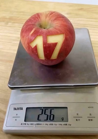 图片中展示了一个红苹果，其表面被雕刻出数字“17”的形状，苹果被放置在一台数字厨房秤上。秤的显示屏清晰显示重量为“256g”，秤的品牌标识“TANITA”和“KOEI”位于秤面下方。苹果整体呈红黄色，果核部分为淡黄色，背景是浅色木质桌面。这张图片通过实物创意的方式，幽默呼应了“苹果17 256g”的说法，将数字与重量结合形成趣味表达。