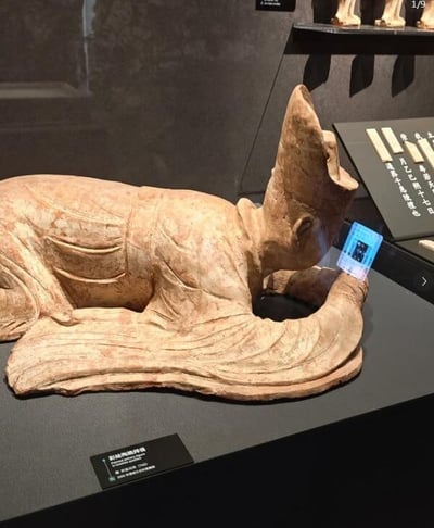 这是一张恶搞梗图，展示了中国古代陶俑在博物馆中展出，雕像手中被PS合成了一部智能手机，看起来就像在低头玩手机，形成明显的时代对比和幽默反差。古老的文士形象和现代手机文化结合，形成‘穿越’感，调侃了现代社会普遍的“低头族”现象。