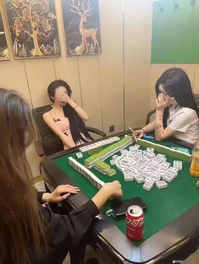 三位女生围坐在麻将桌边，麻将牌杂乱无章地堆在桌子中央，已经无视正常的麻将规则，局面极其混乱，似乎已经完全不是在打麻将了。桌面上还有可口可乐和橙色饮料，场面既搞笑又无奈，非常具有社交网络上的“离谱”气质。这种摆烂局面和女生的轻松姿态形成反差，十分适合表达对聚会娱乐局的吐槽和自嘲。没有出现双关或谐音梗。