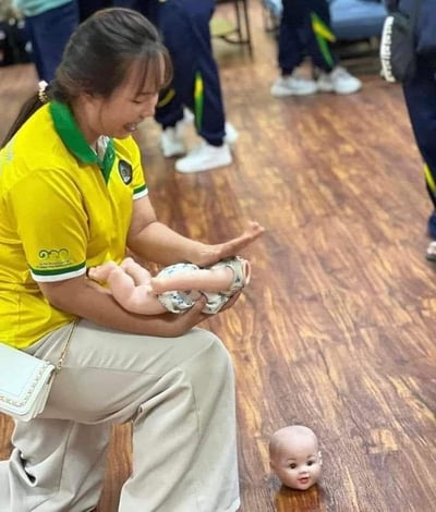 图中一位身着黄绿色上衣、卡其色长裤的女士蹲在室内木质地板上，双手抱着一个假娃娃的身体部分，而地面上的假娃娃头部因为拍摄角度的巧妙错位，看起来像是和身体完全分离，形成了“身首异处”的惊悚又滑稽的视觉效果。女士面带笑容，似乎正在参与一场育儿相关的培训演示，背景中还有几位穿着统一服装的人。这是完全由拍摄角度造成的视觉错觉，并非娃娃真的损坏。
