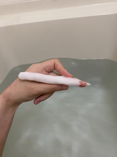 这是一张实拍的静态图片，背景为盛有浅灰色温水的浴缸，画面左侧伸出一只真人的手，手指捏着一长条成型的白色沐浴泡沫，泡沫形态规整修长，外观和白色粉笔高度相似，是泡澡时偶然形成的趣味巧合场景。