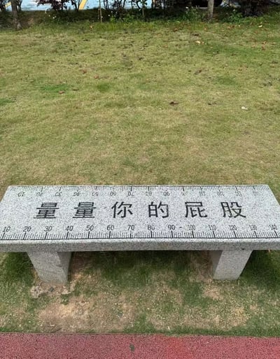 图片中是一张位于草地中的石凳，凳面设计成尺子样式，刻度从10厘米延伸至140厘米，中间用黑色字体写着“量量你的屁股”的趣味文字。石凳两侧有方形支撑腿，背景是修剪整齐的草坪和远处的绿植，整体场景轻松有趣，充满互动感。