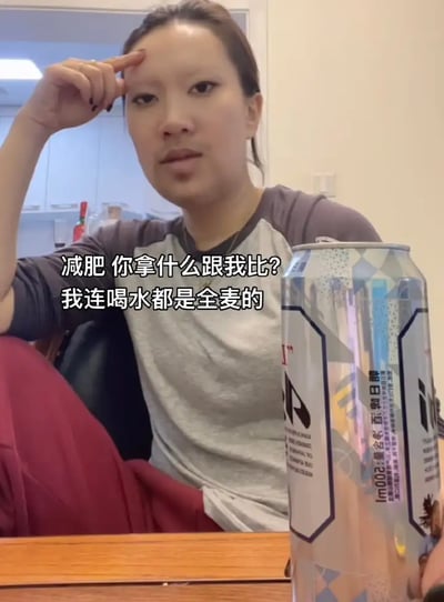 画面中一位女性坐在桌前，桌上放着一罐标注有“全麦”字样的饮品（实际为啤酒或者类似麦芽饮料）。配文：“减肥你拿什么跟我比？我连喝水都是全麦的”，这句话是自嘲减肥的努力以幽默方式表达，暗指“全麦”更健康，但实际上喝的是酒而非水，形成了明显的反差和调侃。

此处的“双关”，既调侃了自律减肥人群爱宣称自己清淡、健康饮食（喝全麦、吃全麦），又通过将“喝水”与“全麦”（啤酒/麦芽饮料）混淆，营造了自欺欺人式的幽默效果。