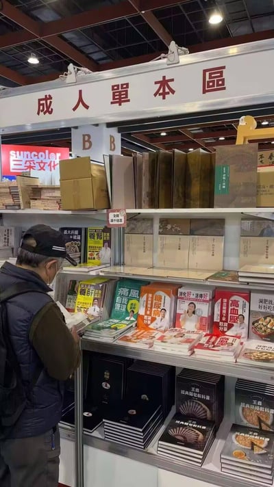 图片展示了一个书展或书店内的场景，货架上方悬挂着醒目的红色标牌“成人单本区”。一位戴着黑色帽子和口罩的老年男性正站在货架前专注地浏览书籍。货架上摆放着多种书籍，主要以健康养生、饮食调理类为主，可见书名包括《糖尿病饮食》《高血压》《健康食谱》等。背景中还能看到其他展位和堆放的纸箱，整体呈现出典型的图书展销会氛围。
