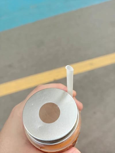 这是一张生活实拍静态图，画面里一只手握着装有橙色饮品的玻璃杯，杯口配有带圆形专用吸管孔的金属螺旋盖，但白色吸管没有穿过预留的孔洞，反而插在了金属盖的外侧边缘，完全无法接触到杯内饮品，呈现出十分荒谬的错误使用状态，背景是带有黄色标线和蓝色边缘区域的灰色户外路面。