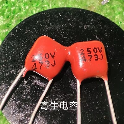 这是一张原始拍摄的静态单图，画面核心是两个连为一体的红色薄膜电容，其外形组合后酷似蝴蝶结，颇具趣味。电容表面印有黑色的参数标识“250V 473J”，其中250V代表电容的额定耐压值为250伏特，473是电容容量的数码标注法，意为47×10³皮法，即47纳法拉，J代表该电容的容量误差等级为±5%。图片底部配有白色的说明文字“寄生电容”，电容放置在带有细微毛絮的黑色平面上，背景是绿色的纹理材质。这类薄膜电容属于电子电路中常用的被动元件，可用于滤波、信号耦合等场景，而“寄生电容”通常指电路中意外产生的电容效应，此处也可能是对两个电容连接形态的趣味化标注。