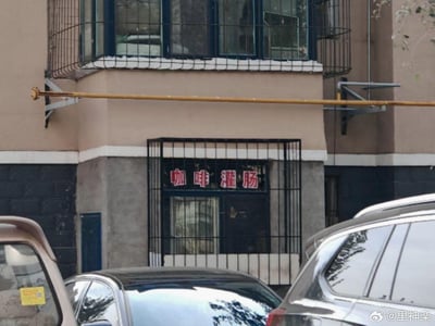 这是一张实拍的居民楼照片，楼体采用上下分层的配色设计，底层的窗户装有黑色防盗栏，窗户上方贴着红底白字的「咖啡灌肠」字样。咖啡作为饮品和灌肠的医疗行为结合，形成了极具违和感的迷惑组合，让人觉得滑稽有趣。画面下方可见几辆停放的汽车，楼体外部还有管道、防护栏等居民楼常见设施。
