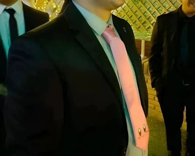 一张在正式场合拍摄的照片，中心人物穿着黑色西装、浅蓝色衬衫，系着一条粉色领带。特别引人注目的是领带上画着卡通风格的笑脸，有两只大眼睛和腮红，看起来像某种可爱的卡通角色。背景中还有其他穿着西装的人，环境装饰有格子结构和串灯，营造出正式而温馨的氛围。
