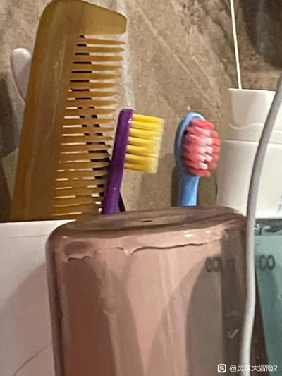 这是一张浴室场景的实拍图，紫色手柄黄刷头、蓝色手柄粉刷头的牙刷被放置在浅棕色容器后方，因视觉错位，牙刷刷头刚好与容器边缘重合，看起来像是容器“长”出了类似人脸的模样，旁边还立着一把木梳，整体画面充满趣味，是利用视角巧合形成的搞笑视觉错觉。