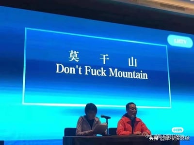 这是一张会议现场的实拍图，画面中背景的蓝色大屏幕上显示着中文地名“莫干山”，但下方的英文翻译却被恶搞成了低俗的“Don't Fuck Mountain”，利用“莫干”的发音做了恶意谐音梗，制造出荒诞又尴尬的搞笑效果。画面下方有两位真实人物，左侧的人低头看着手中的文件，神情专注；右侧的人穿着红色外套，对着麦克风，似乎正在发言。整个场景结合错误又低俗的翻译，形成强烈的反差感，成为网络上流传的搞笑梗图。