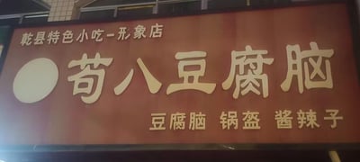 这是一张实拍的线下小吃店招牌图，招牌底色为红棕色，文字采用醒目的米白色。招牌最上方标注“乾县特色小吃-形象店”，明确了店铺的地域特色定位；中间是大号字体的核心店名“苟八豆腐脑”，左侧搭配一个圆形标识，突出店铺主打产品；下方则清晰列出了店铺的主营餐品：豆腐脑、锅盔、酱辣子，都是陕西乾县的经典特色美食。整体设计简洁朴实，信息传递直接，能让过往食客快速了解店铺的品类和特色。