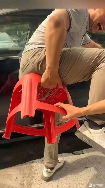一名身穿灰色短袖和卡其色裤子的男子正坐在红色塑料凳上，身体前倾，嘴巴大张，表情夸张地大笑。另一只手从下方扶住凳子，背景中可见黑色汽车，推测为户外街道或停车场场景。画面充满喜剧效果，展现了日常生活中的搞笑瞬间。