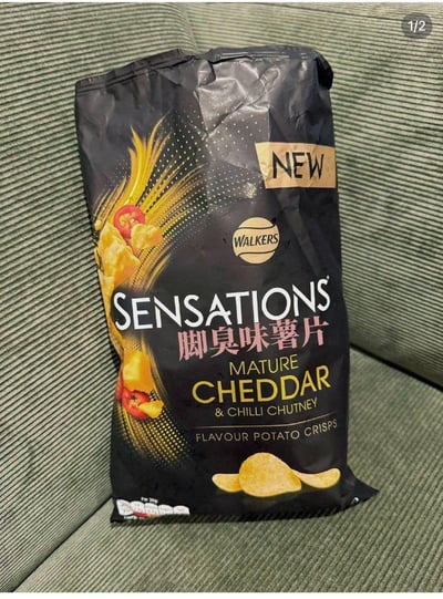 图片中展示了一袋黑色包装的Walkers Sensations薯片，放置在绿色条纹沙发上。包装袋上原本的“MATURE CHEDDAR & CHILLI CHUTNEY”（成熟切达奶酪与辣椒酸辣酱）口味被幽默地修改为中文“脚臭味薯片”，形成强烈的反差萌。金色的薯片图案和红色辣椒点缀在包装上，顶部标有“NEW”字样，整体设计夸张又搞笑，显然是网友通过图片编辑软件制作的趣味改图，旨在用奇葩口味引发笑点。