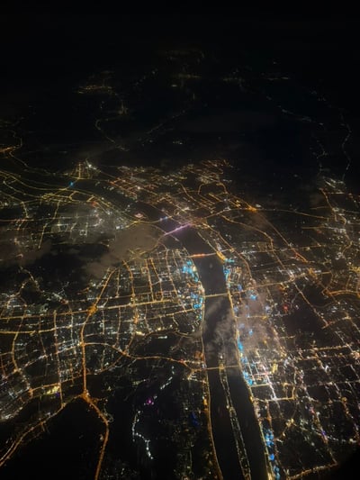 这是一张夜间从高空（大概率为民航飞机舷窗视角）拍摄的城市全景图，一条宽阔的河流纵向穿城而过，河流上可见多座亮灯的跨江桥梁连接两岸；城市整体路网呈规整的网格布局，暖黄色的道路照明灯光与建筑的白、蓝、紫等各色灯光交织，清晰勾勒出城市建成区的范围与形态，画面中还漂浮着少量轻薄云雾。发布者最初误以为该城市是武汉，对比特征后发现不符，遂发起猜城市的互动。
